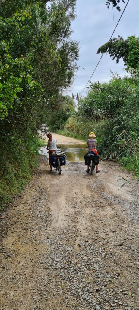 2 vakantiefietsers staan op een gravelweg voor een beek die dwars over de weg loopt. Aan weerszijden van de weg is hoge, groene begroeiing.